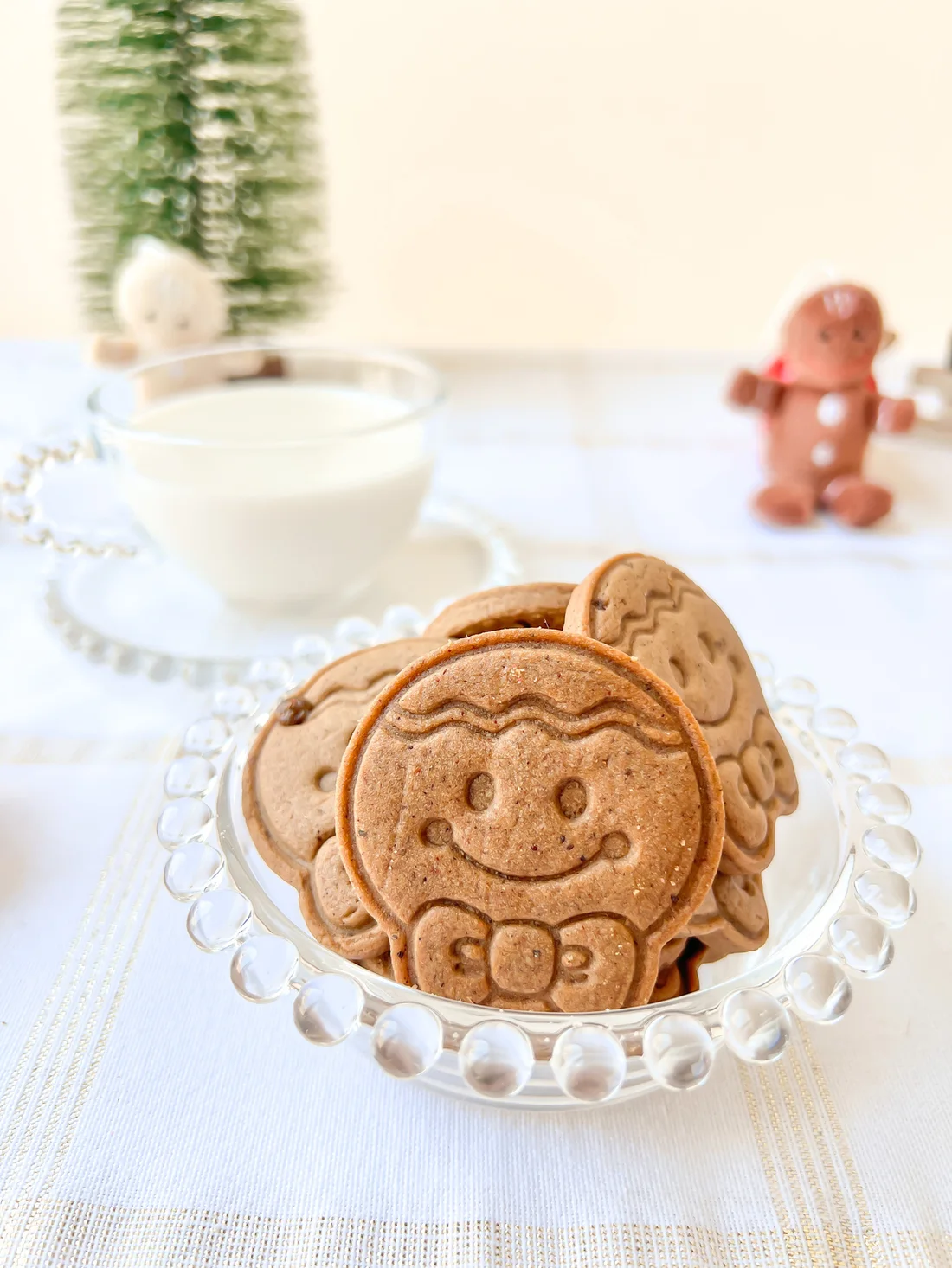 Prato de vidro com biscoitos de gingerbread com carinha, xícara de leite e pinheiro ao fundo